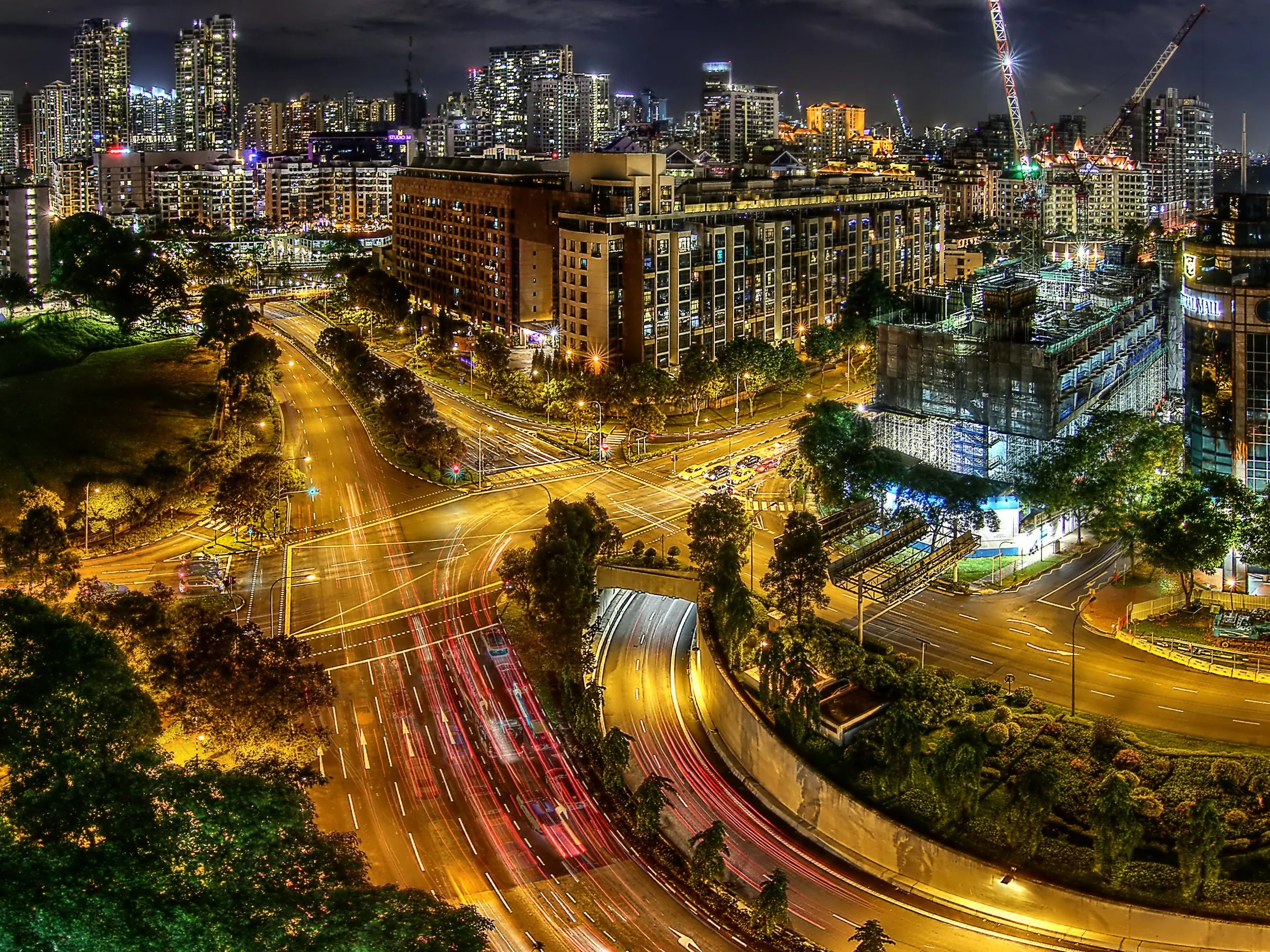 Night view over River Valley from above, with roads curving past condominium blocks and a wooded slope in the foreground and residential towers along the skyline
