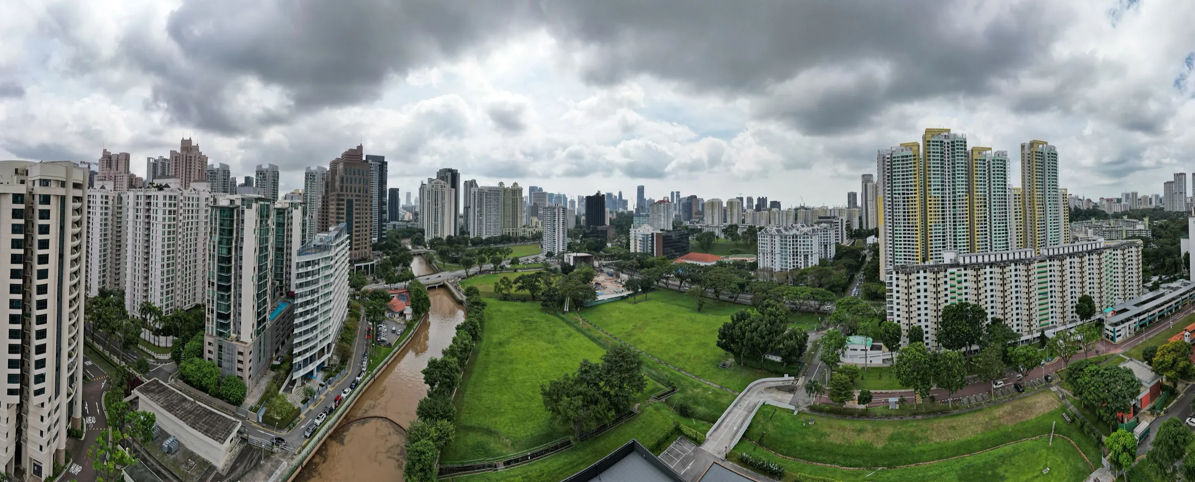 Aerial panorama of the Singapore River in River Valley, the canal running between condominium towers on the left and open green fields and HDB blocks on the right, with the city skyline beyond