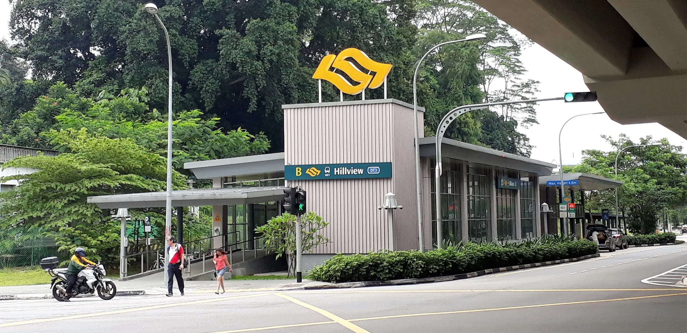 Exit B of Hillview MRT station, a low glass-walled building with a tall pillar bearing the MRT logo and Hillview DT3 sign, set against dense trees at a road junction
