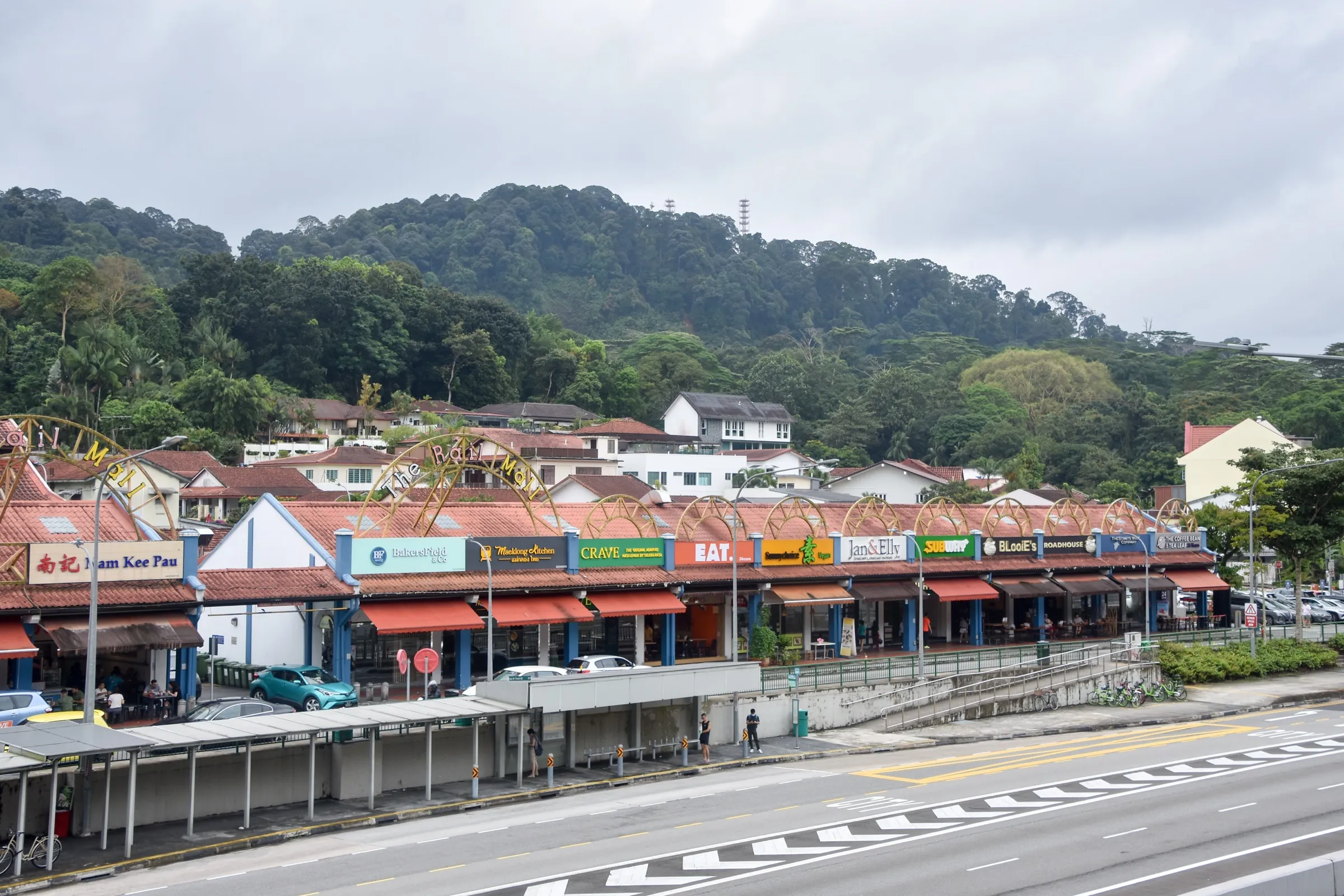 The Rail Mall on Upper Bukit Timah Road, a single-storey row of shops and restaurants under red-tiled roofs and decorative arches, with houses and the forested slopes of Bukit Timah Hill behind