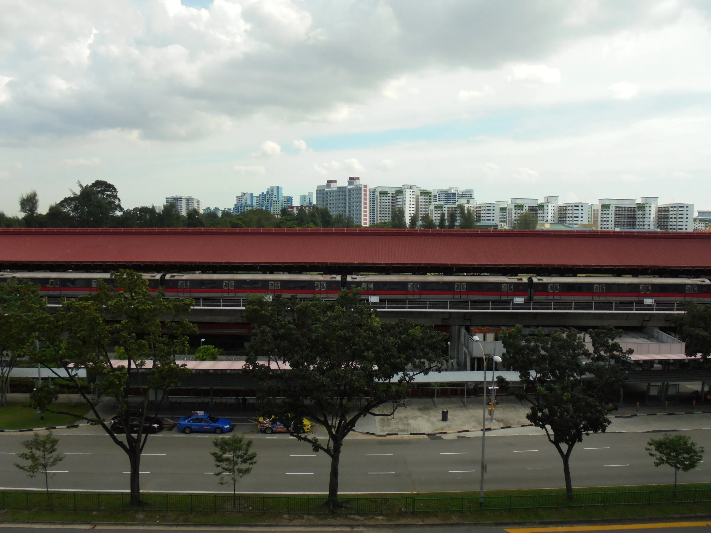 Lakeside MRT station seen from across the road, an elevated platform under a long red roof with a train at the platform and HDB blocks on the skyline